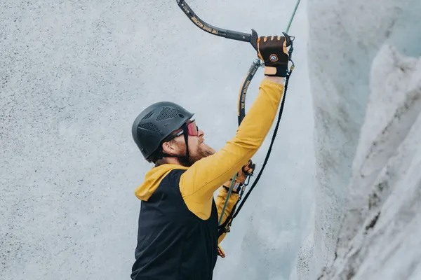 A client in a yellow jacket and black helmet holds his ice axe, about to take a swing at the base of a white glacial wall and begin the ice climb.