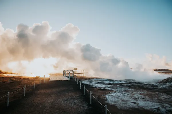 Geothermal steam billows, creating puffy white clouds that float over a walkway as the sun begins to set.