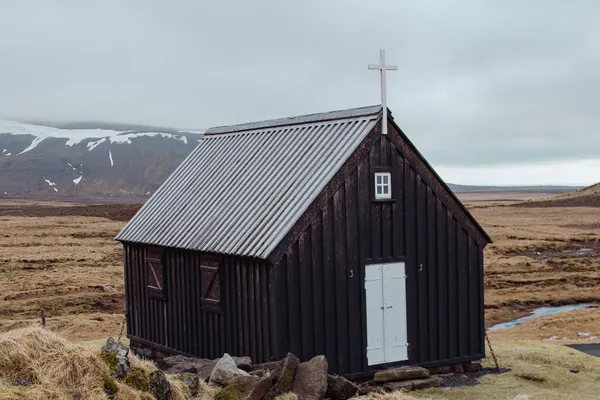 A small, old church constructed of dark wood sits on a light yellow meadow with lightly snow covered mountains in the background on an autumn day.