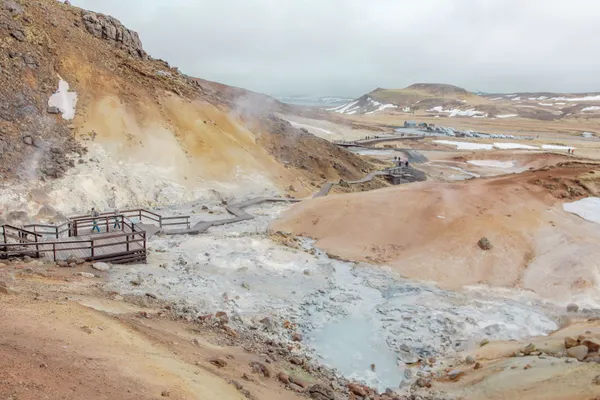 Tourists on a long, wooden walkway observe geothermal steam rising up from light- grey muddy puddles surrounded by tan, peach and rust-colored hilly terrain.
