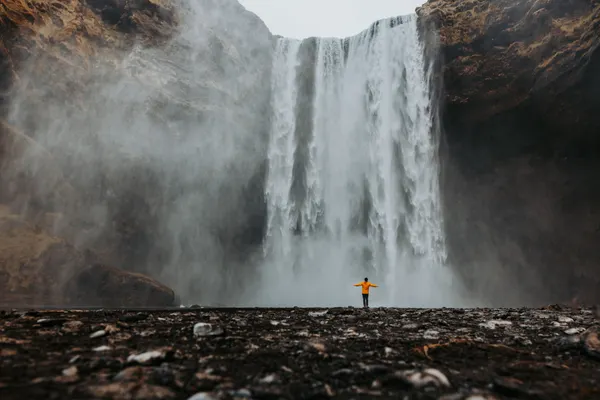 A person in an orange jacket holds up their arms welcoming the spray of Iceland's massive Skógafoss.