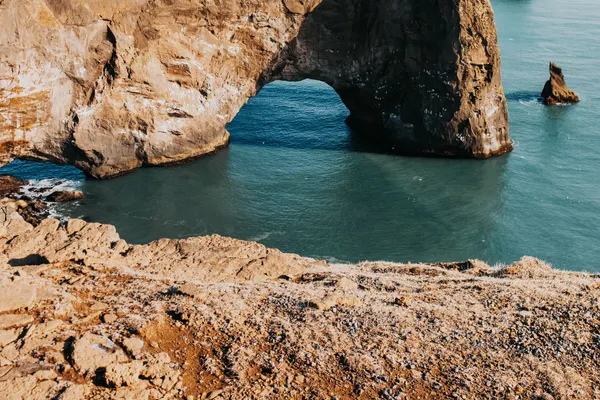 A promontory juts out into the ocean, ending with a massive rock arch on a sunny day.