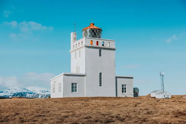 The white rectangular lighthouse with an orange dome at Dyrhólaey viewpoint, with Mýrdalsjökull ice cap in the background on a clear day.