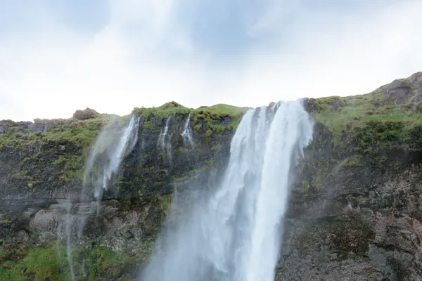 A waterfall blows sideways in Iceland's strong wind on a summer day, with bright green grass in the foreground.