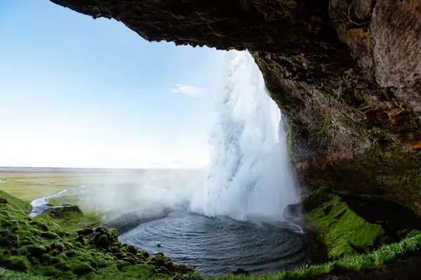 View from the back of Seljalandsfoss pouring into a small pool as it sprays mist, partially concealing the green field in the background.