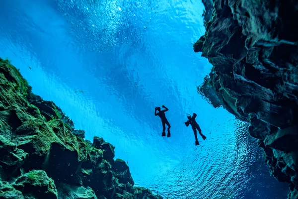 Sillouettes of two freedivers up on the surface of Silfra's vibrant blue waters between rocky fissure walls.