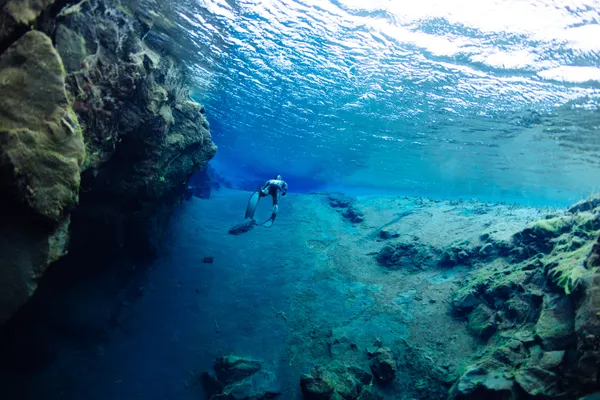 Sun sparkles on the surface of the water, as a free diver explores below along a fissure wall and sandy bottom.