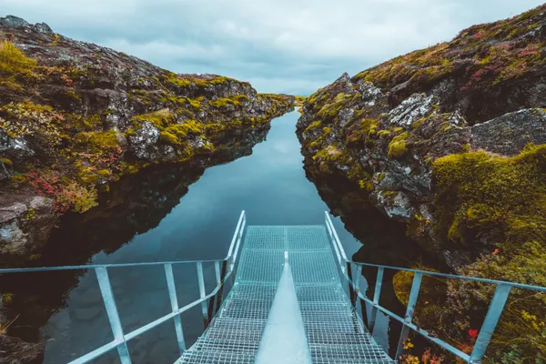 Metal stairs lead into the narrow Sifra fissure framed by moss- covered rocky walls on each side and the water is very still, reflecting the cloudy skies above.