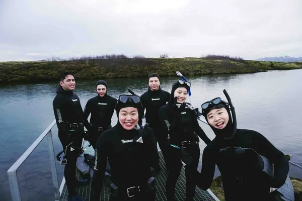 A group of happy tourists wearing wetsuits, dive masks and snorkels stand smiling in front of Silfra after the free diving tour.
