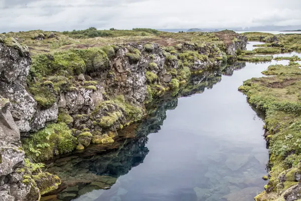 A long, narrow water- filled rift surrounded by moss covered rocks reflects sun on a partially cloudy day
