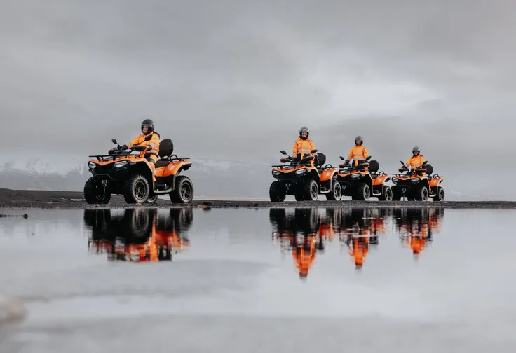 The reflections of four people on ATVs are seen in a small, still pond as they ride past on a cloudy day.