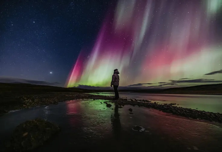 An aurora gazer stands on a path of rocks in a small pond, enjoying the colorful northern lights above.