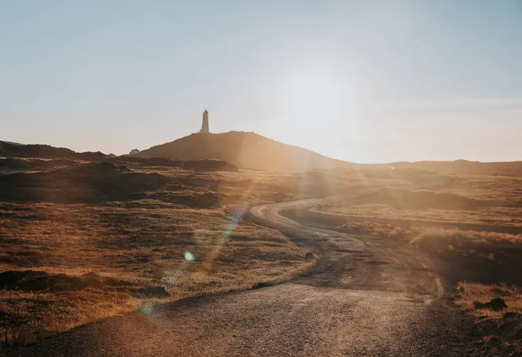 A dirt road curves up an orange hill, leading to a lighthouse as the sun shines brightly casting sun spots. 