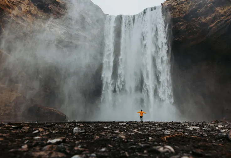 A person in an orange jacket holds up their arms welcoming the spray of Iceland's massive Skógafoss.