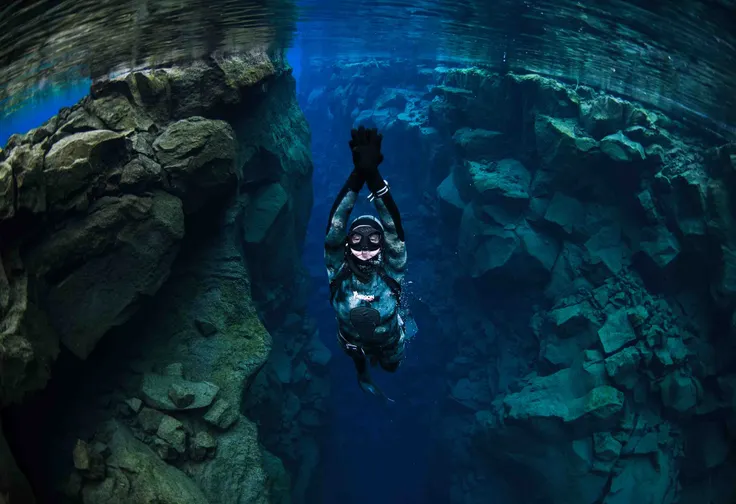 A freediver with hands together swimming through bright blue water towards the surface.
