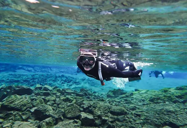 A snorkeler smiling under the clear water just below the surface, explores a shallow part of silfra.