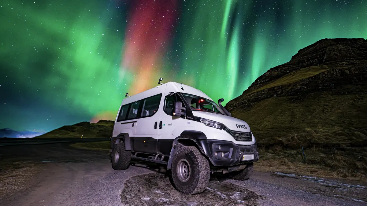 On tour, the 4x4 vehicle called the Super Truck is parked next to a small grassy mountain while the Northern lights look like a colorful explosion in the sky.
