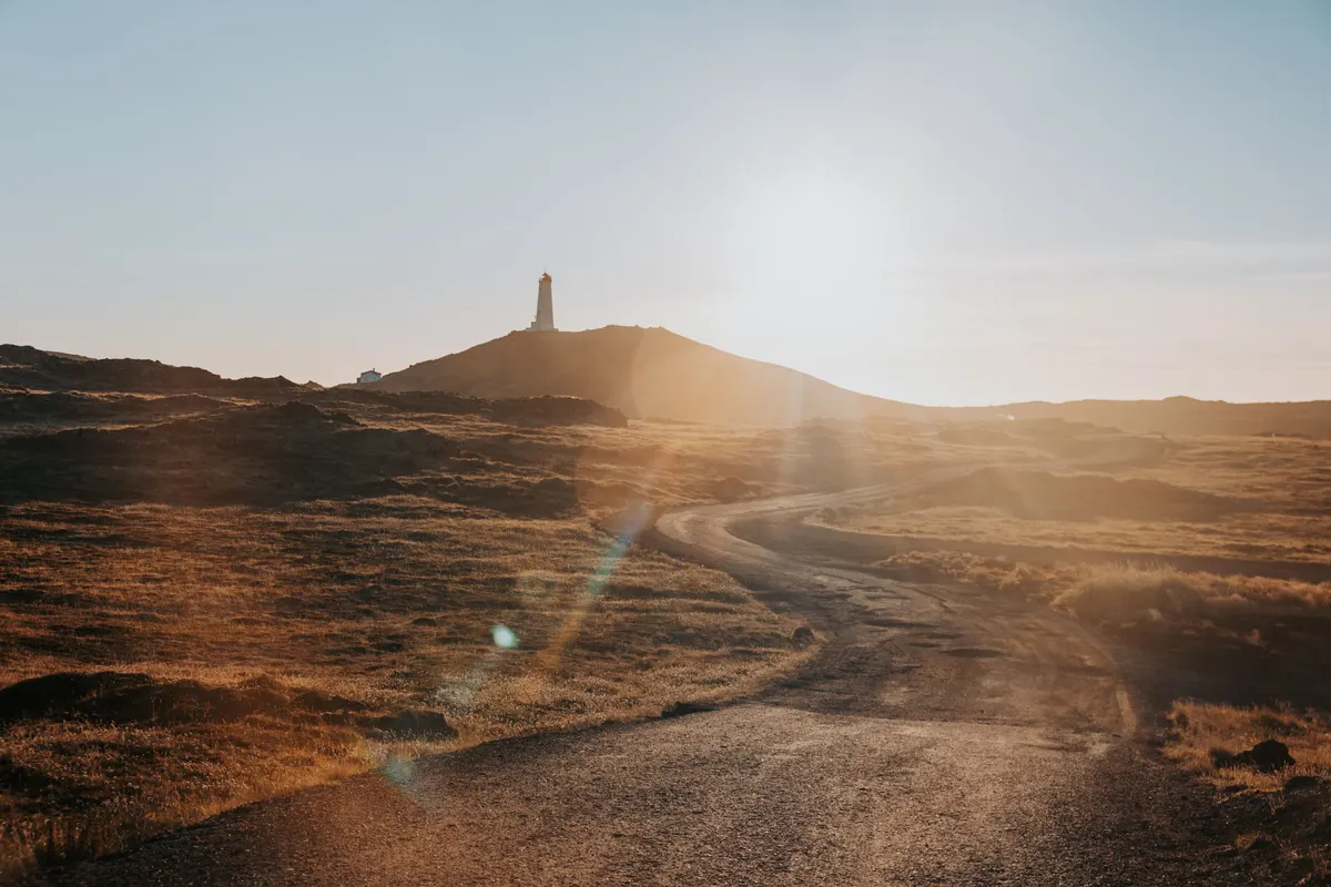 A dirt road curves up an orange hill, leading to a lighthouse as the sun shines brightly casting sun spots. 