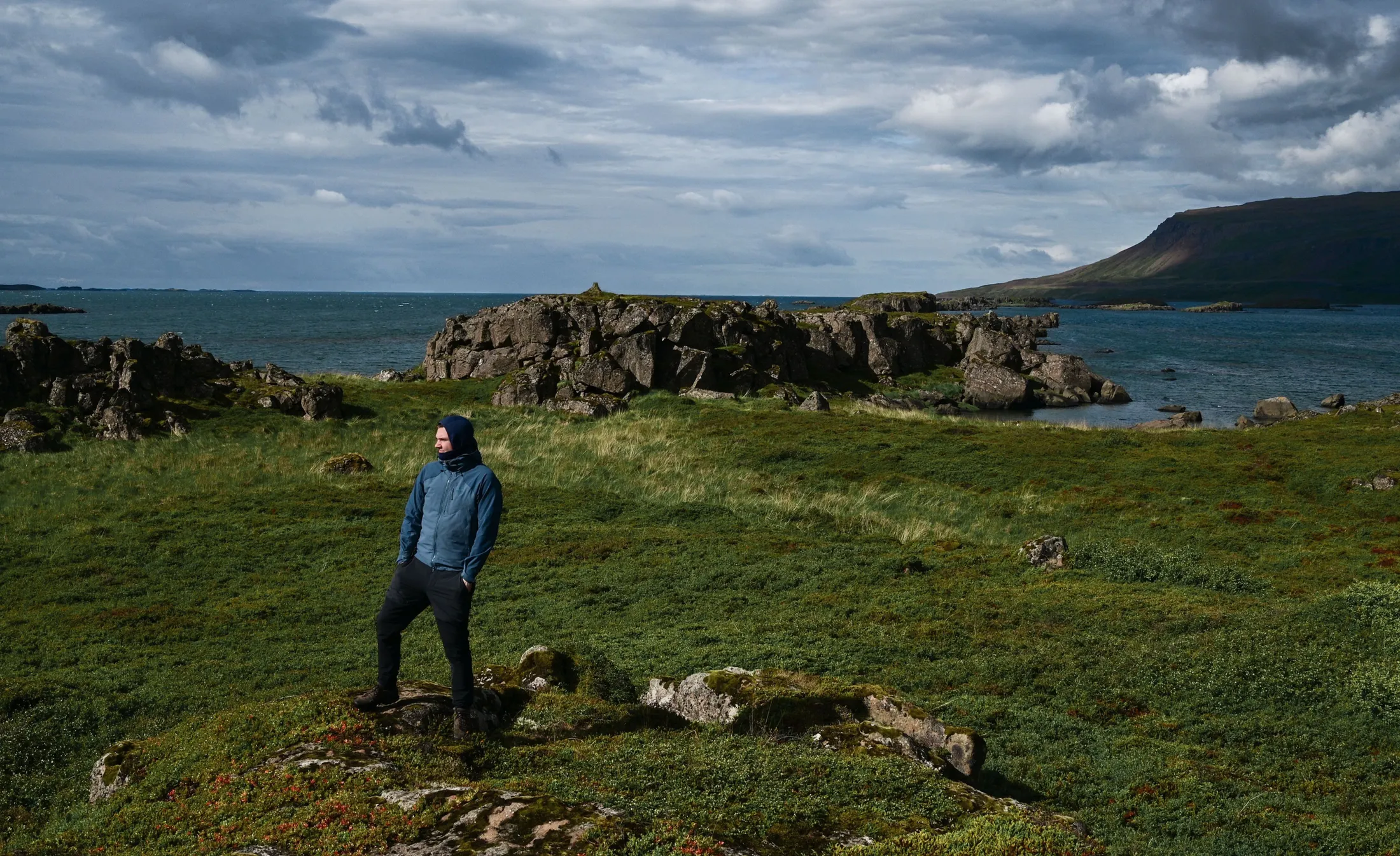 Man in a blue softshell jacket stands in a grassy area next to a fjord on a sunny, windy day.