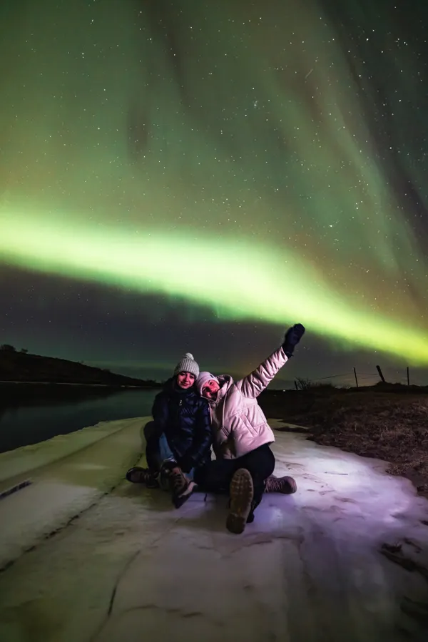 Two people sit on the icy ground below a sky lit up by a light green aurora.