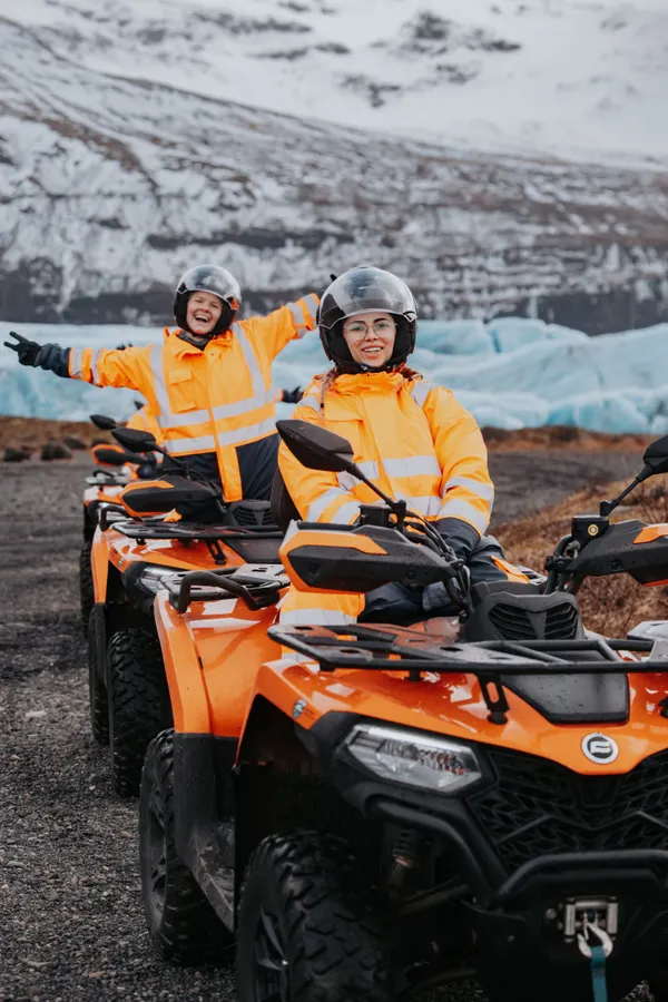 Two ATV riders in bright orange coveralls pose excitedly in front of a vibrant blue glacial outlet in winter.
