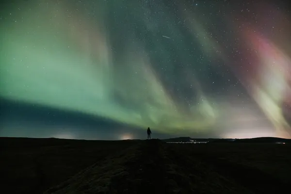 A person's silhouette appears very small under a sky completely covered with light green Northern lights.