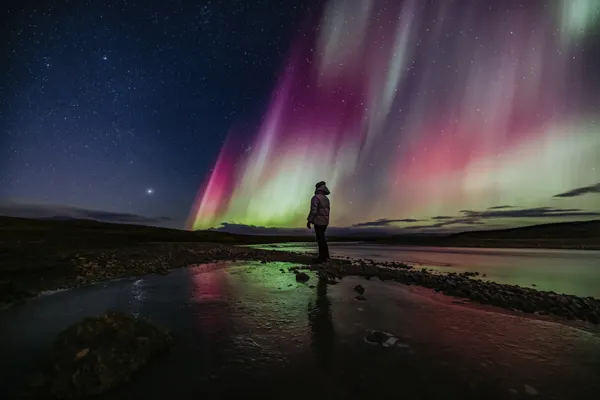 An aurora gazer stands on a path of rocks in a small pond, enjoying the colorful northern lights above.