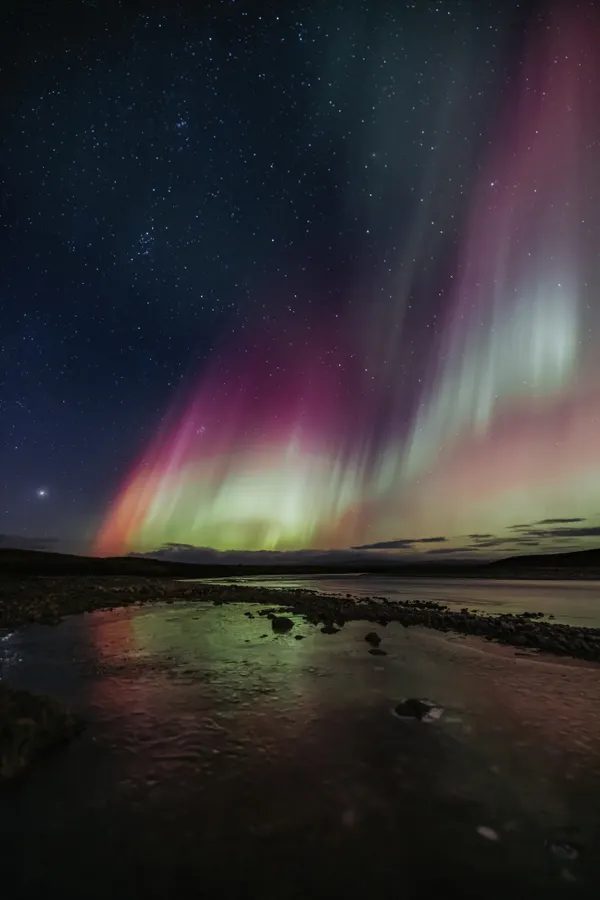 A faint glow of green and purple northern lights are reflected in a stream as they dance above in the dark sky.