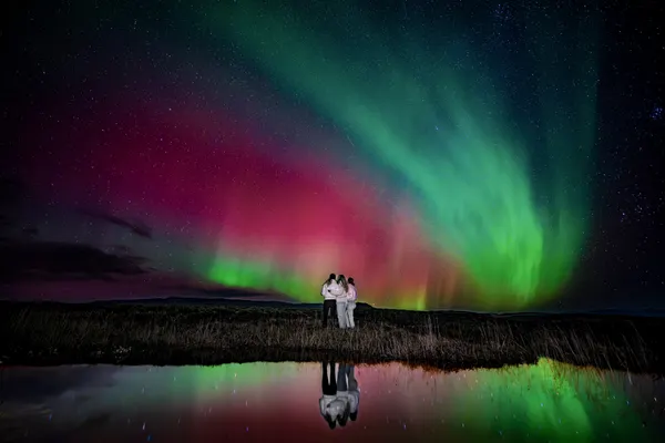 Three people on a small group Northern Lights tour stand on the edge of a pond under a vibrant aurora.