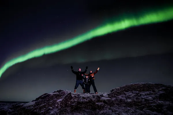A family of three on a Northern Lights tour stand on a snowy hill with their arms up under a single vibrant green aurora line.
