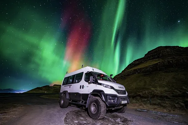 On tour, the 4x4 vehicle called the Super Truck is parked next to a small grassy mountain while the Northern lights look like a colorful explosion in the sky.