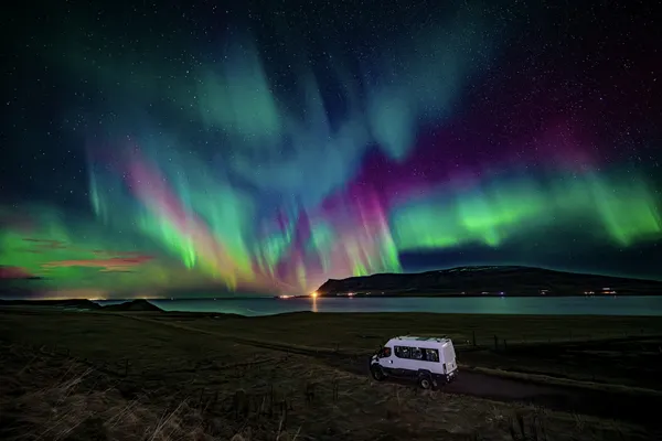 The 4x4 Super truck parks next to a lake, while very colorful northern lights explode across the sky.