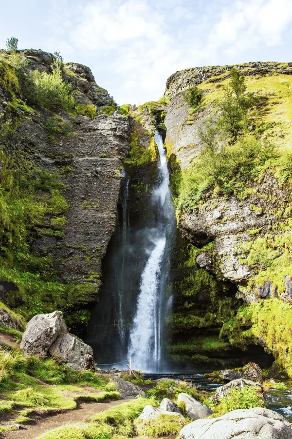 A partially concealed waterfall flows between layered rock formations covered in green moss on a sunny day.