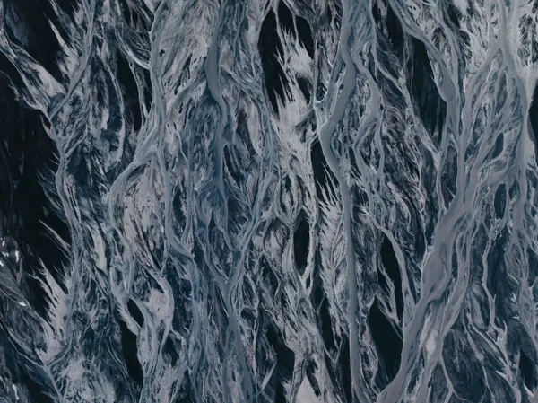 Hundreds of small, light blue glacial river streams flow over deep black volcanic sand in a unique weave-like pattern.