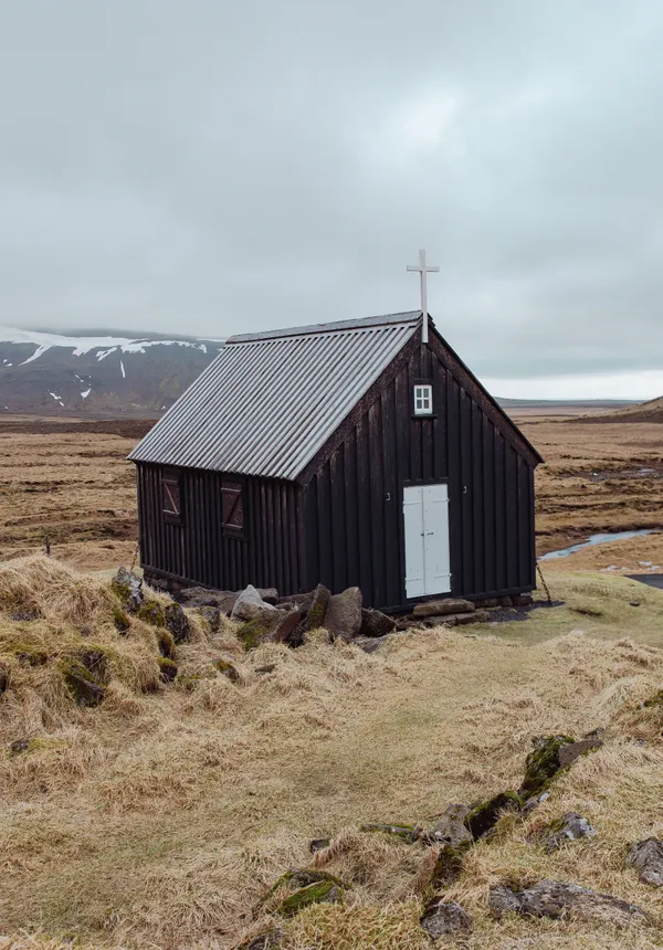 A small, old church constructed of dark wood sits on a light yellow meadow with lightly snow covered mountains in the background on an autumn day.