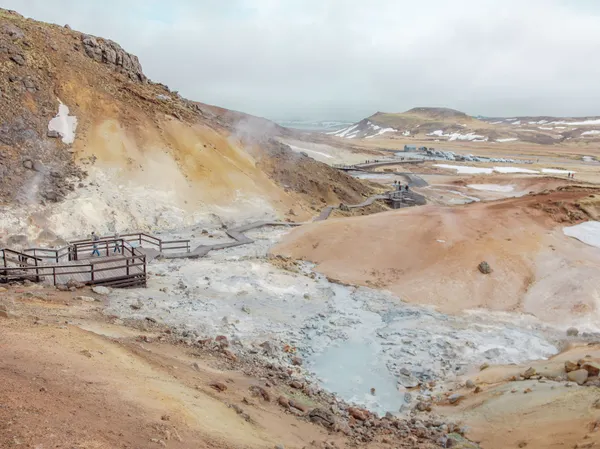 Tourists on a long, wooden walkway observe geothermal steam rising up from light- grey muddy puddles surrounded by tan, peach and rust-colored hilly terrain.