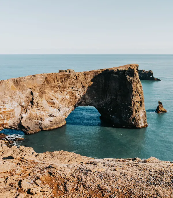 A promontory juts out into the ocean, ending with a massive rock arch on a sunny day.