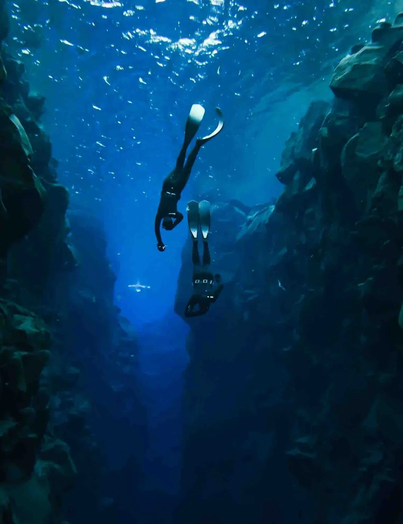 Two freedivers dive deeper into a fissure filled with clear, deep blue water.