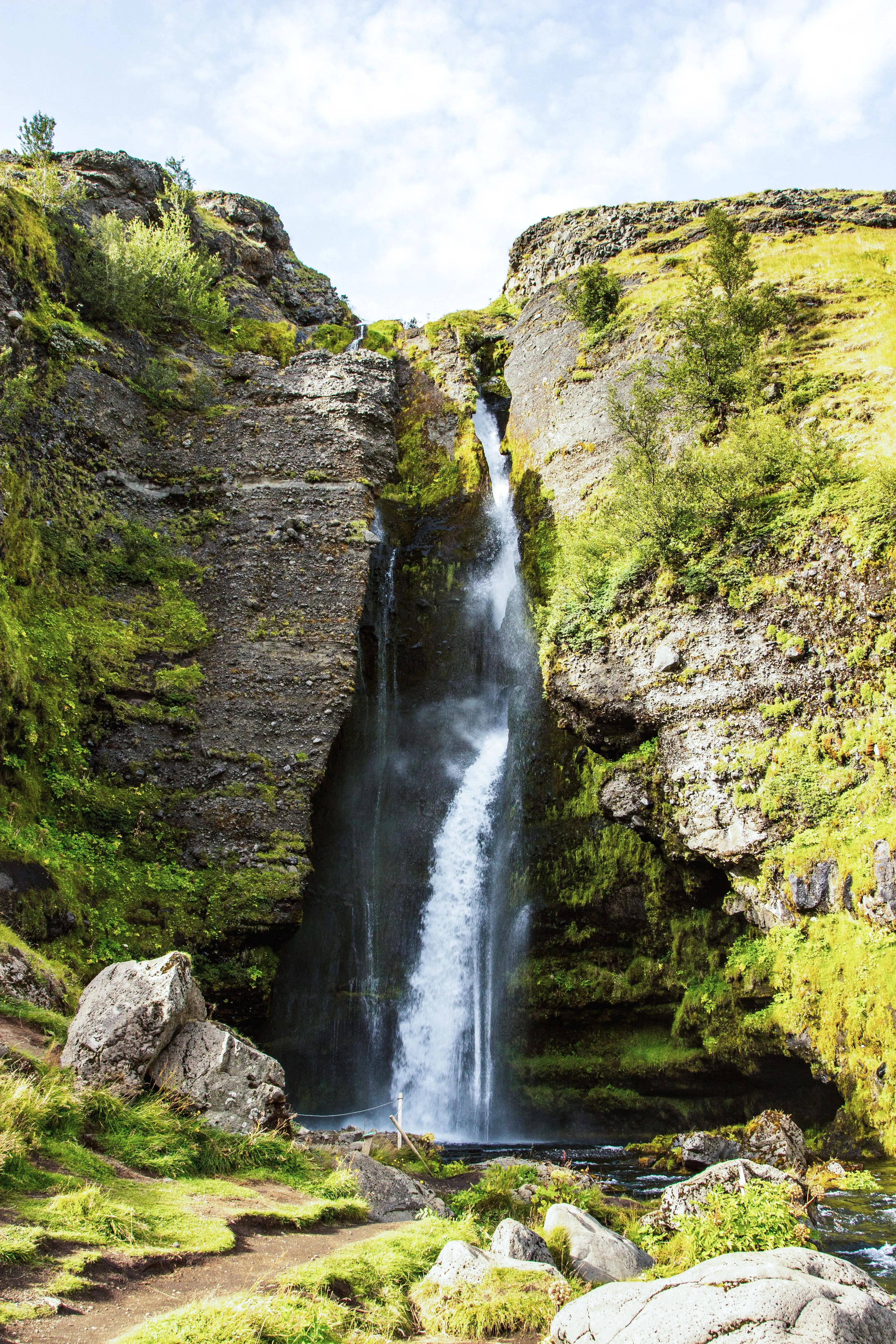 A partially concealed waterfall flows between layered rock formations covered in green moss on a sunny day.