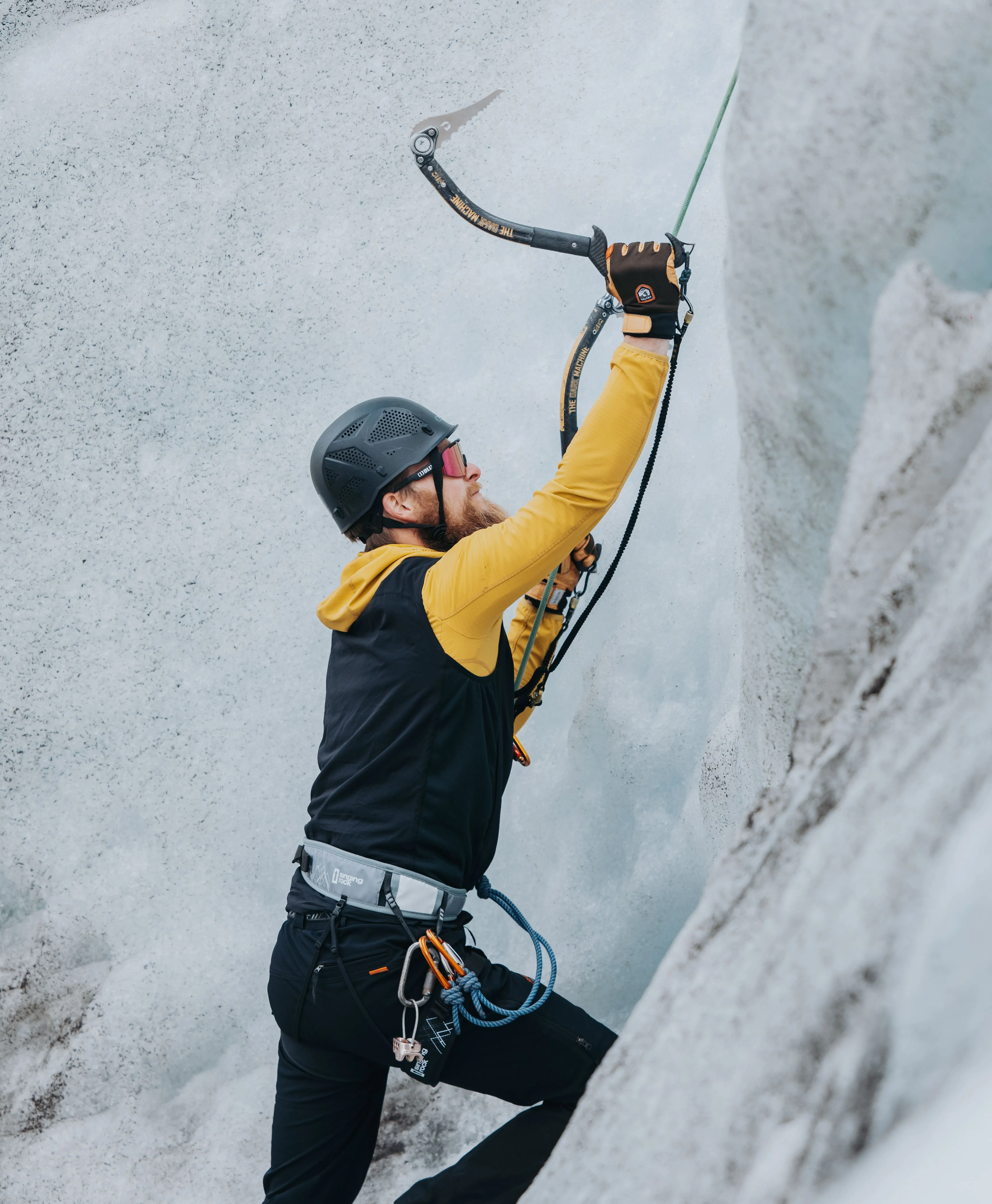A client in a yellow jacket and black helmet holds his ice axe, about to take a swing at the base of a white glacial wall and begin the ice climb.