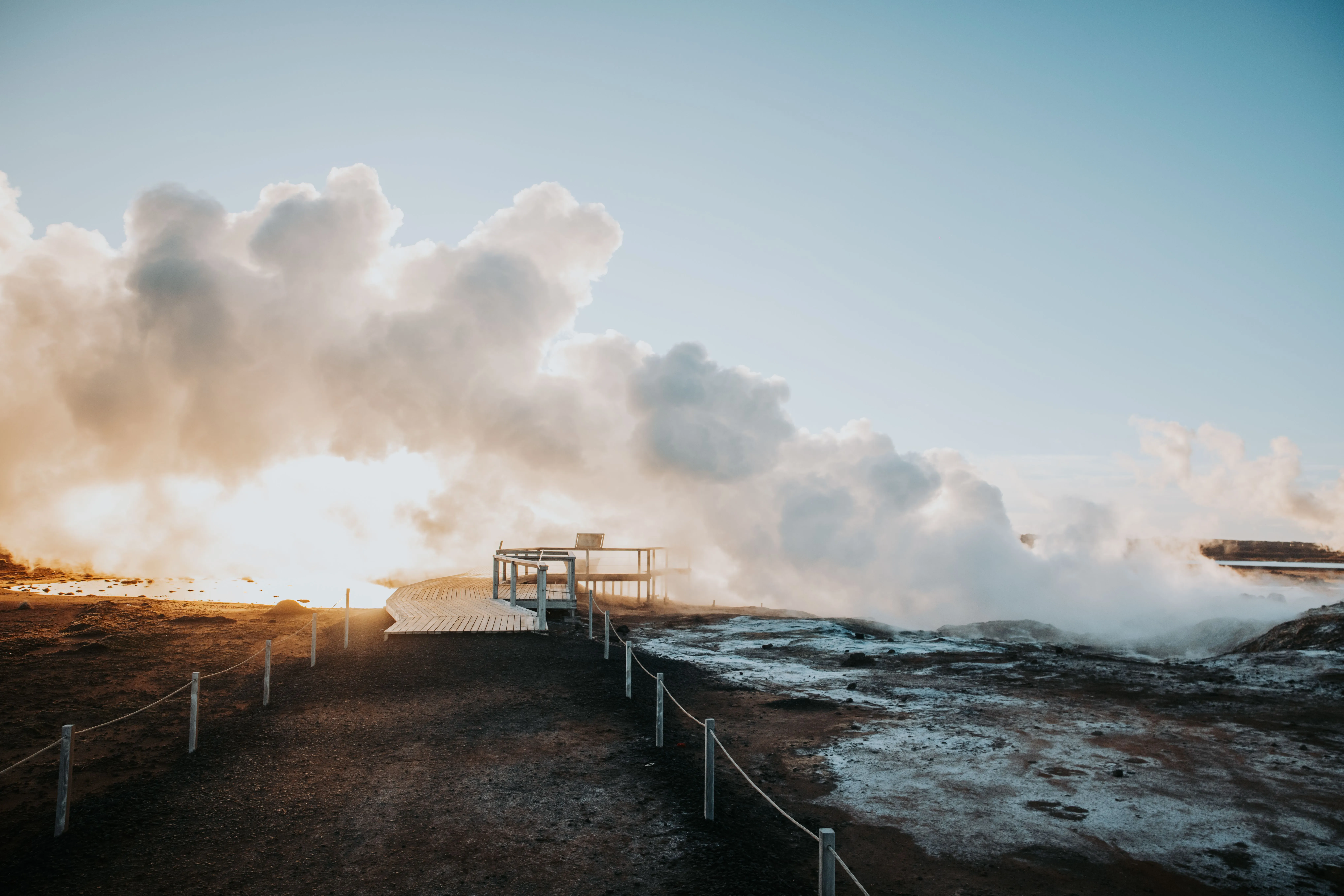 Geothermal steam billows, creating puffy white clouds that float over a walkway as the sun begins to set.