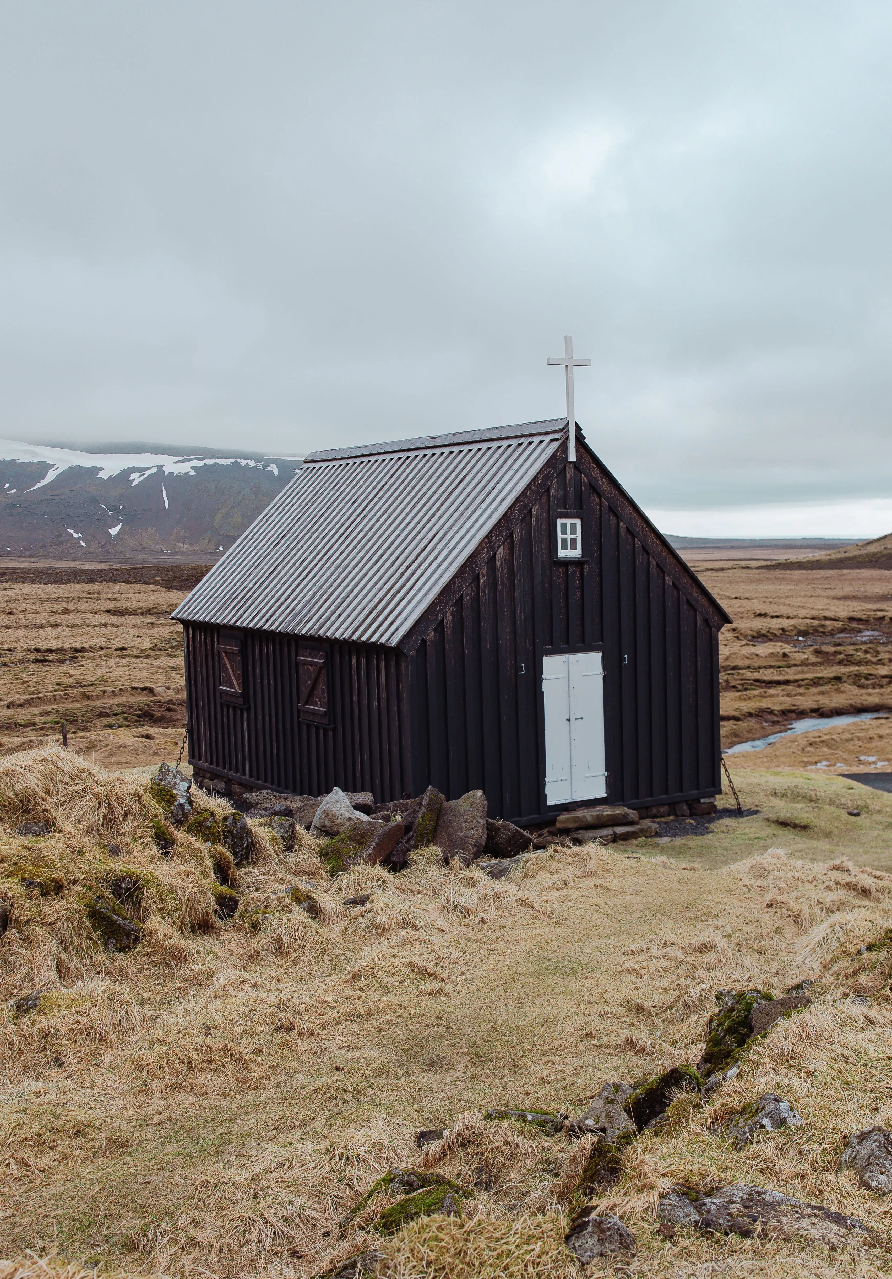 A small, old church constructed of dark wood sits on a light yellow meadow with lightly snow covered mountains in the background on an autumn day.