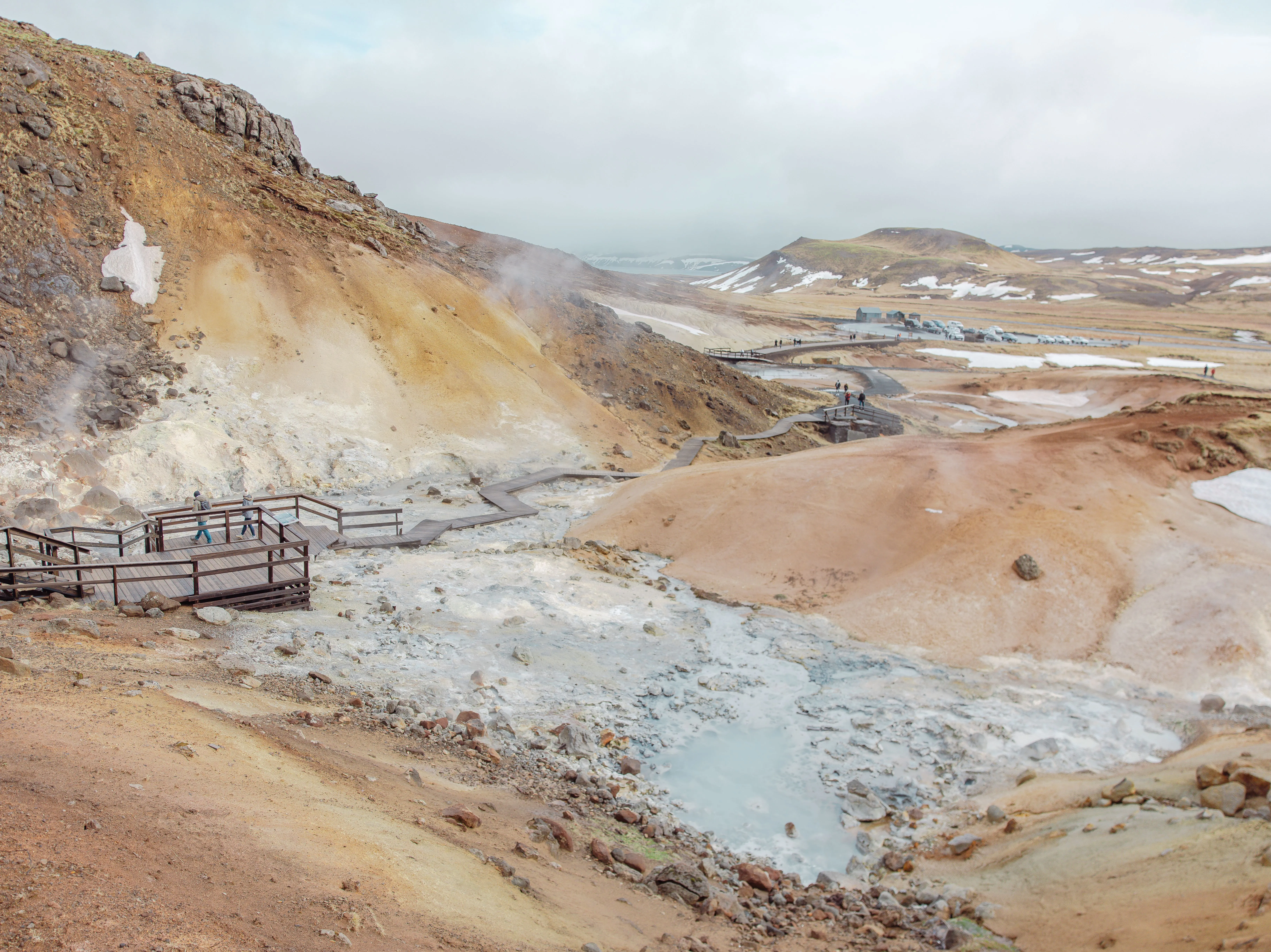 Tourists on a long, wooden walkway observe geothermal steam rising up from light- grey muddy puddles surrounded by tan, peach and rust-colored hilly terrain.