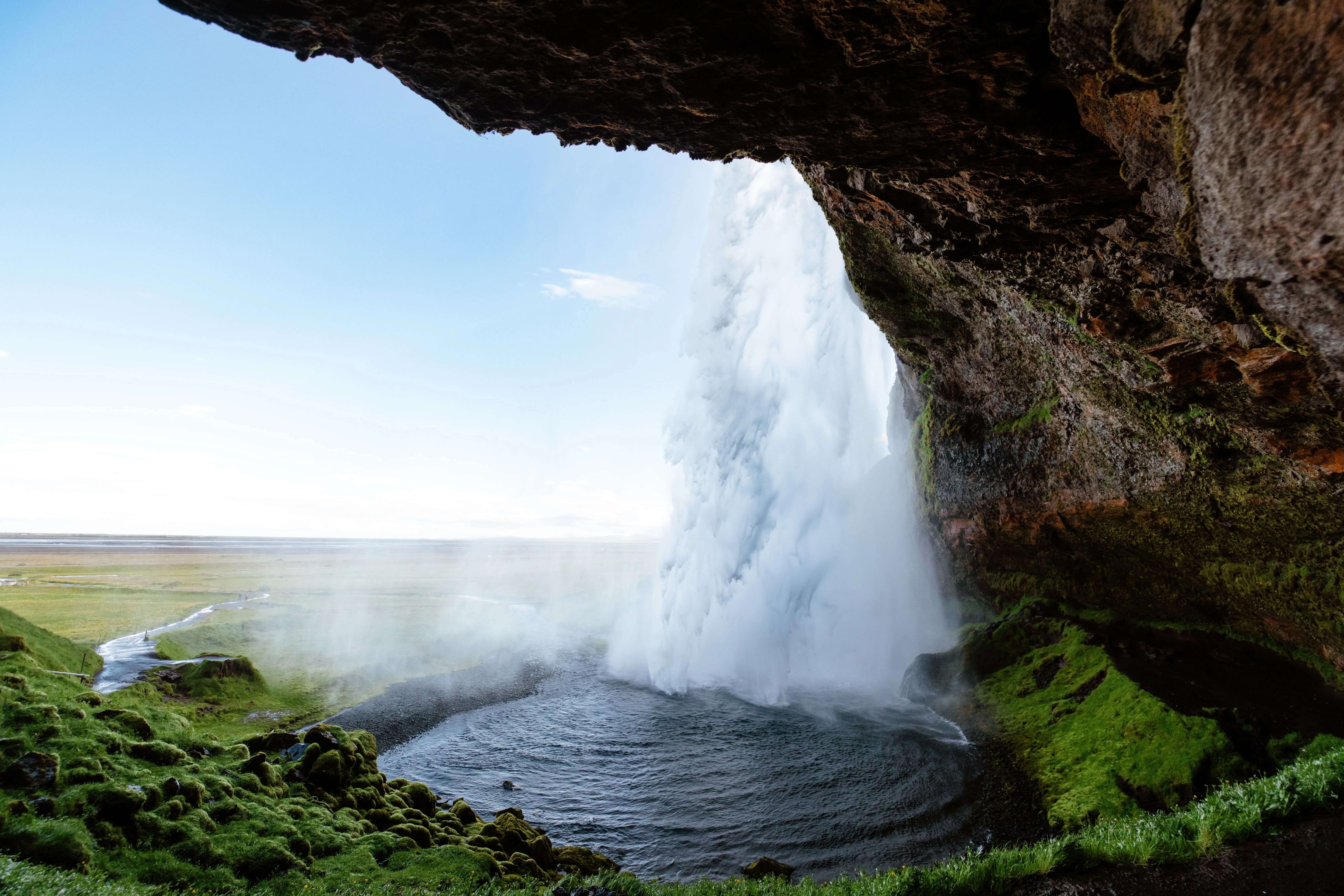 View from the back of Seljalandsfoss pouring into a small pool as it sprays mist, partially concealing the green field in the background.