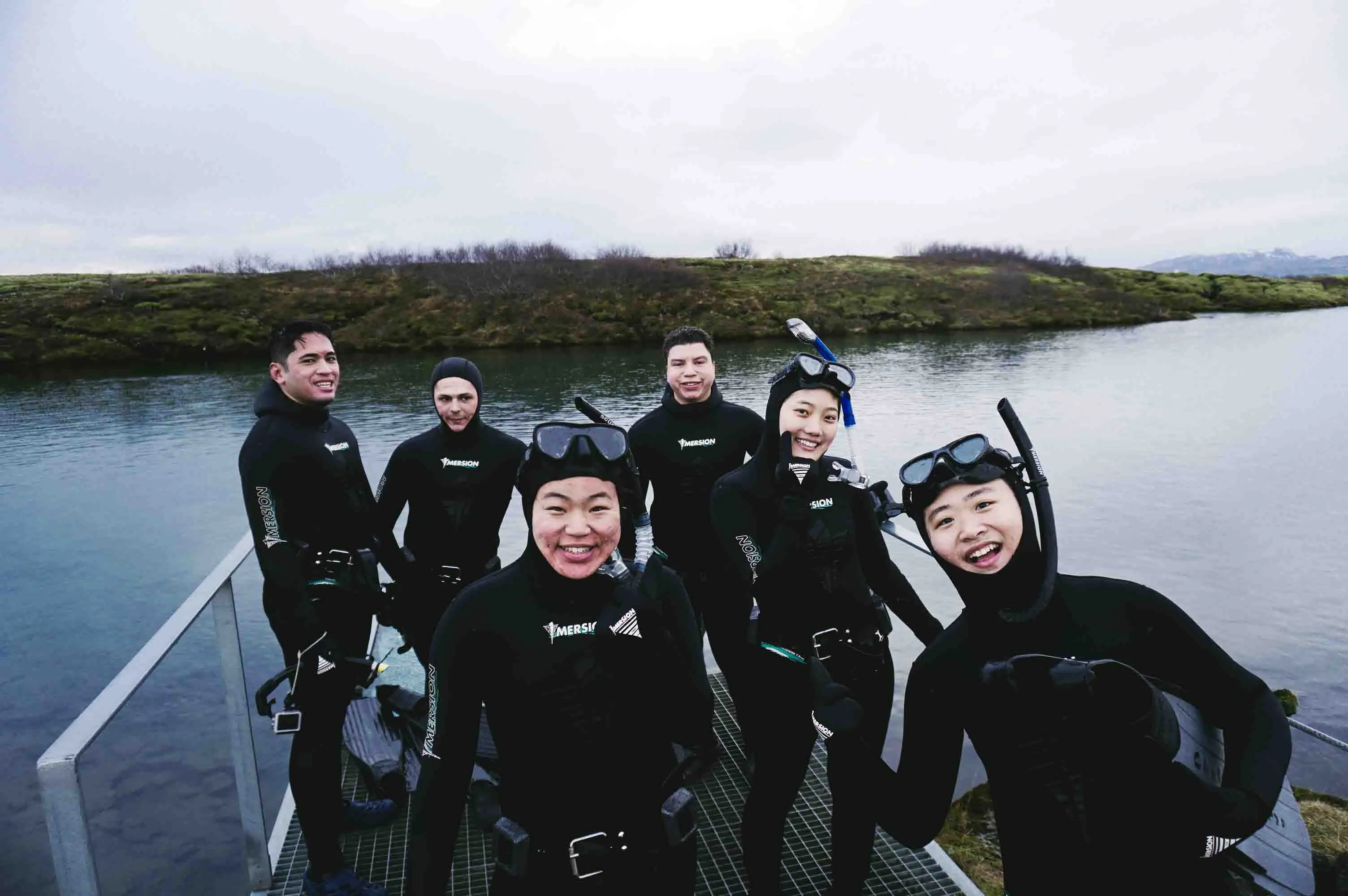 A group of happy tourists wearing wetsuits, dive masks and snorkels stand smiling in front of Silfra after the free diving tour.