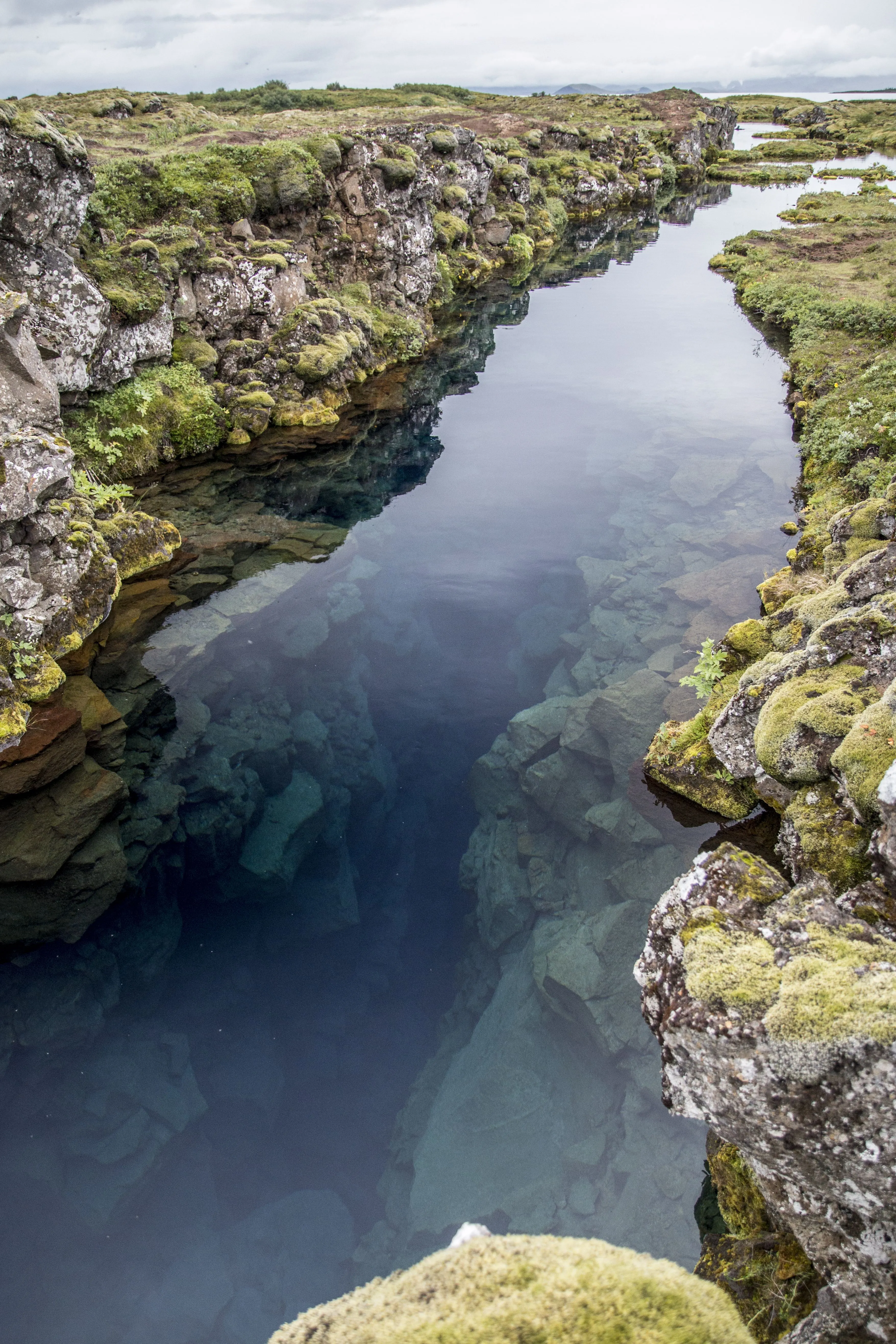 A long, narrow water- filled rift surrounded by moss covered rocks reflects sun on a partially cloudy day