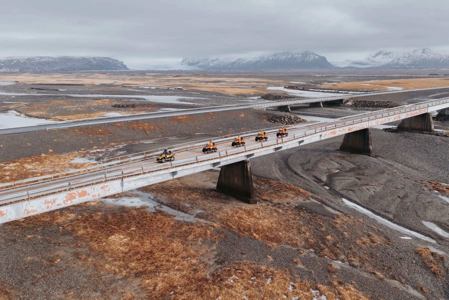 Riders on ATVs cross a long bridge over a dried up glacial river bed with glacier covered mountains in the background